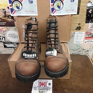 May include: A pair of brown leather Grinders boots with black laces and soles. The boots have a classic design with a round toe and are displayed with tags that read "Made in England". The boots are on a cardboard box.
