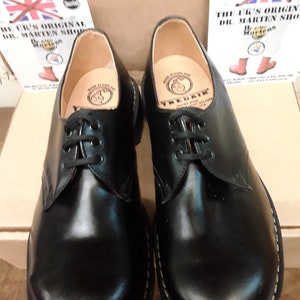 May include: A pair of black leather lace-up shoes with a white stitching detail around the sole. The shoes have a brown leather interior with a circular logo that reads "TREADAIR MADE IN ENGLAND".