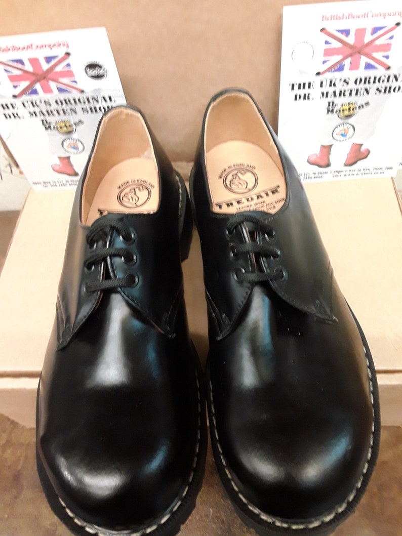 May include: A pair of black leather lace-up shoes with a white stitching detail around the sole. The shoes have a brown leather interior with a circular logo that reads "TREADAIR MADE IN ENGLAND".