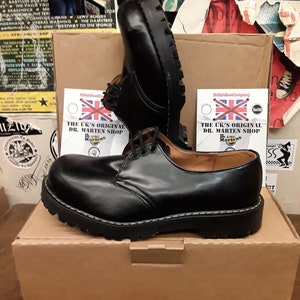 May include: A pair of black leather Dr. Martens shoes with a lace-up design and a thick, black sole. The shoes are sitting on a brown cardboard box with a white label that reads "The UK's Original Dr. Martens Shop".