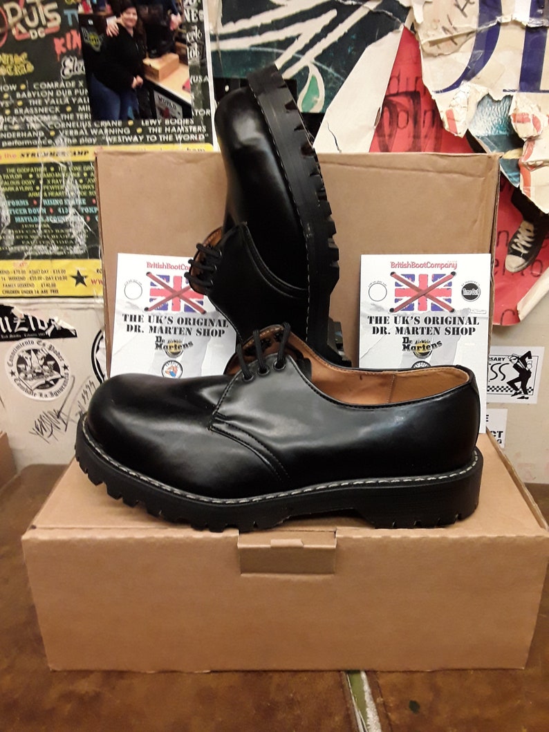 May include: A pair of black leather Dr. Martens shoes with a lace-up design and a thick, black sole. The shoes are sitting on a brown cardboard box with a white label that reads "The UK's Original Dr. Martens Shop".