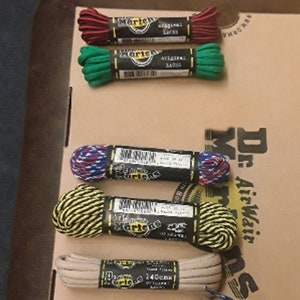 May include: Six pairs of Dr. Martens shoelaces in various colors and patterns, including red, green, blue, yellow, and beige. The laces are packaged in small cardboard boxes with the Dr. Martens logo.