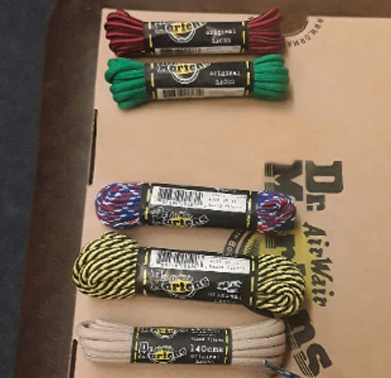 May include: Six pairs of Dr. Martens shoelaces in various colors and patterns, including red, green, blue, yellow, and beige. The laces are packaged in small cardboard boxes with the Dr. Martens logo.