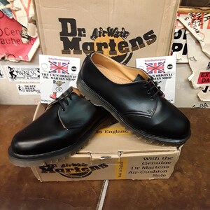 以下が含まれることがあります： 黒色のDr. Martensの靴。靴紐付きで、ブランドロゴ入りの段ボール箱の上に置かれています。靴は革製のアッパーと耐久性のあるソールを備えたクラシックなデザインです。