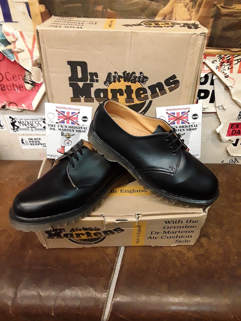 以下が含まれることがあります： 黒色のDr. Martensの靴。靴紐付きで、ブランドロゴ入りの段ボール箱の上に置かれています。靴は革製のアッパーと耐久性のあるソールを備えたクラシックなデザインです。