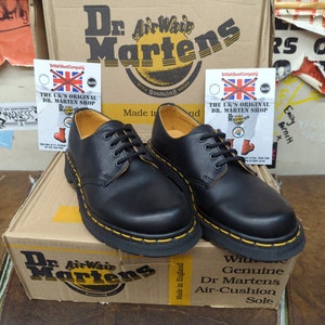 May include: A pair of black leather Dr. Martens boots with yellow stitching and a yellow air-cushioned sole. The boots are sitting on a box with the Dr. Martens logo and the text "Made in England".