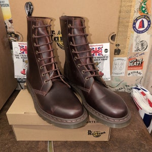 May include: A pair of brown leather Dr. Martens boots with laces and a yellow stitching detail. The boots are on a box with the Dr. Martens logo.