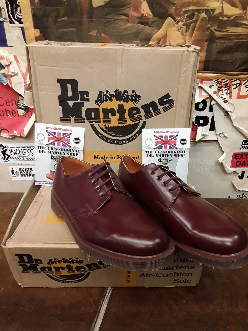 Puede incluir: Un par de zapatos de cuero burdeos Dr. Martens con cordones marrones, exhibidos sobre una caja de cart&oacute;n. La caja presenta el logotipo de Dr. Martens y el texto "AirWair with Bouncing Soles".