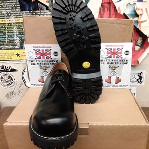 May include: Black leather lace-up shoes with a thick, black rubber sole. The shoes are sitting on a brown cardboard box with a white label that says "The UK's Original Dr. Martens Shop".