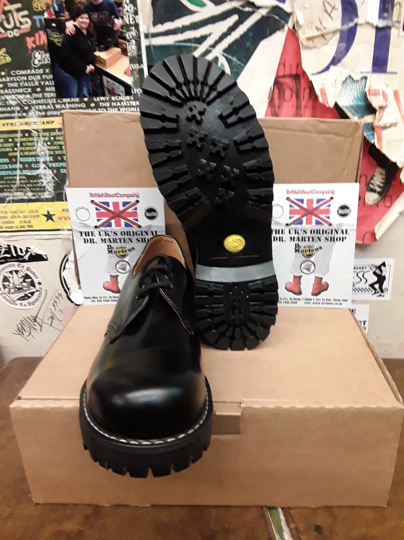 May include: Black leather lace-up shoes with a thick, black rubber sole. The shoes are sitting on a brown cardboard box with a white label that says "The UK's Original Dr. Martens Shop".