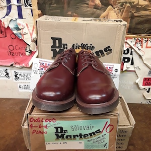 Puede incluir: Un par de zapatos oxblood Dr. Martens Solovair lisos, talla 10, con suela marr&oacute;n, presentados en una caja de cart&oacute;n. La caja muestra el nombre de la marca y los detalles del producto. El fondo incluye carteles y otra caja.