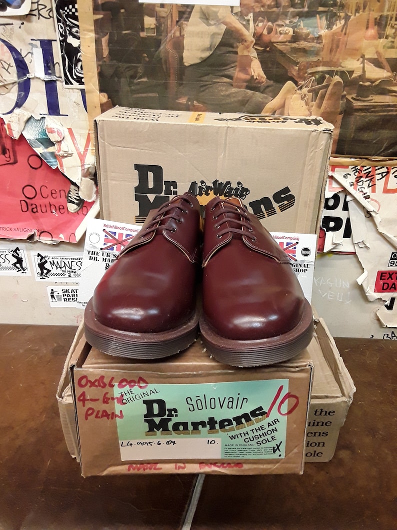 Puede incluir: Un par de zapatos oxblood Dr. Martens Solovair lisos, talla 10, con suela marr&oacute;n, presentados en una caja de cart&oacute;n. La caja muestra el nombre de la marca y los detalles del producto. El fondo incluye carteles y otra caja.