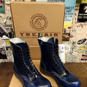 Könnte beinhalten: Ein Paar blaue Lederstiefel mit schwarzen Schnürsenkeln und einer dicken schwarzen Sohle. Die Stiefel sind in England hergestellt und haben eine Goodyear-Rahmenkonstruktion. Die Stiefel stehen auf einem braunen Karton mit den Worten "Treadair" und "Foam-Injected" darauf.