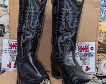 Pode incluir: Um par de botas de cowboy de couro preto com pontas prateadas. As botas têm costuras intrincadas e uma ponta pontiaguda.