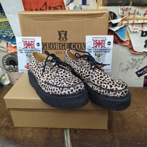 May include: A pair of brown and white leopard print shoes with black laces and soles. The shoes are sitting on a brown box with the text "GEORGE COX EST. 1" printed on it. The box also has a sticker that says "THE UK'S ORIGINAL DR. MARTEN SHOP".