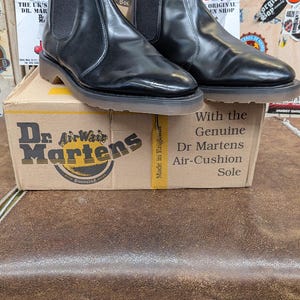 Puede incluir: Un par de botines Chelsea negros Dr. Martens con paneles laterales elásticos, sobre una caja de cartón. La caja presenta el logotipo y el texto de Dr. Martens. Los botines tienen un acabado brillante y un diseño clásico.