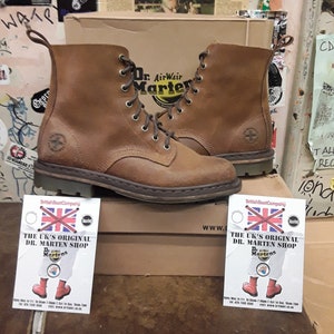 Puede incluir: Un par de botas Dr. Martens de cuero marrón con cordones negros. Las botas están sobre una caja con el texto "The UK's Original Dr. Martens Shop" y el logotipo de Dr. Martens.