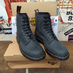 May include: A pair of black leather Dr. Martens boots with yellow stitching. The boots have a snakeskin pattern and are sitting on a brown box with the Dr. Martens logo.