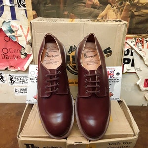 Puede incluir: Un par de zapatos Oxford Dr. Martens burdeos con cordones, presentados en su caja de cart&oacute;n original. Los zapatos tienen un dise&ntilde;o cl&aacute;sico con costuras visibles y una suela robusta. La caja muestra el logotipo y la marca Dr. Martens.