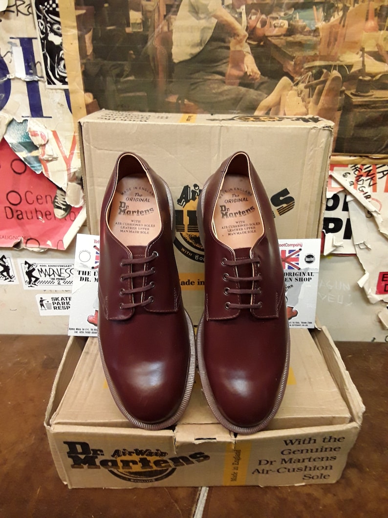 Puede incluir: Un par de zapatos Oxford Dr. Martens burdeos con cordones, presentados en su caja de cart&oacute;n original. Los zapatos tienen un dise&ntilde;o cl&aacute;sico con costuras visibles y una suela robusta. La caja muestra el logotipo y la marca Dr. Martens.