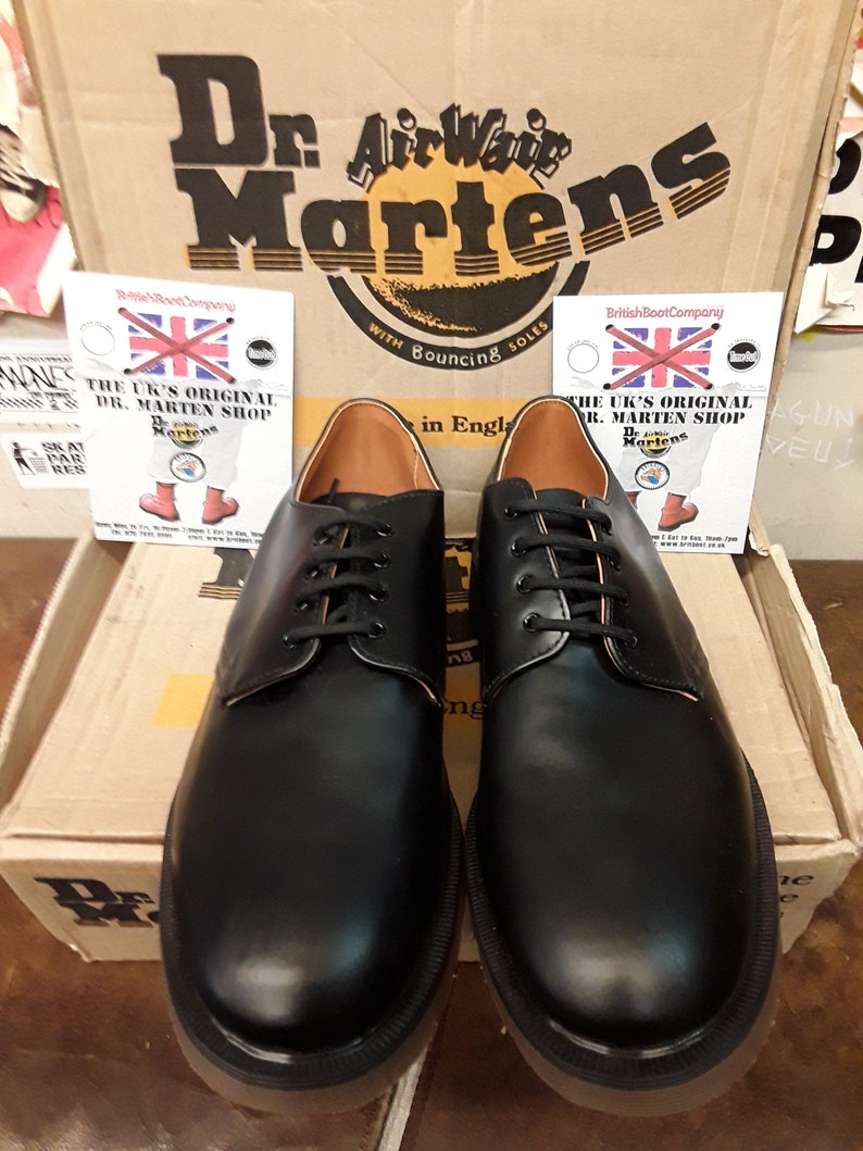 K&ouml;nnte beinhalten: Ein Paar schwarze Lederschuhe von Dr. Martens mit Schn&uuml;rsenkeln und braunem Lederfutter. Die Schuhe stehen auf einem Karton mit dem Dr. Martens Logo und dem Text "With Bouncing Soles".