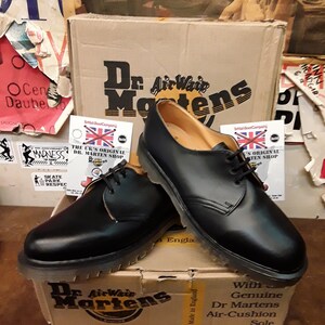 以下が含まれることがあります： 黒色のDr. Martensオックスフォードシューズのペア。靴紐付きで、段ボール箱の上に展示されています。箱にはDr. Martensのロゴとテキストが表示されています。靴は、革のアッパーと耐久性のあるソールを備えたクラシックなデザインです。