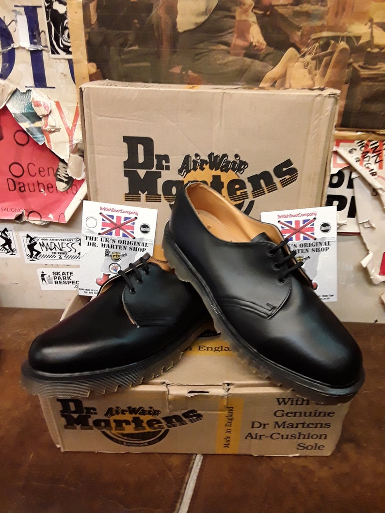 以下が含まれることがあります： 黒色のDr. Martensオックスフォードシューズのペア。靴紐付きで、段ボール箱の上に展示されています。箱にはDr. Martensのロゴとテキストが表示されています。靴は、革のアッパーと耐久性のあるソールを備えたクラシックなデザインです。