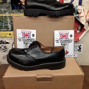 May include: A pair of black leather Dr. Martens shoes with a white stitching detail on the sole. The shoes are sitting on a brown cardboard box with the text "The UK's Original Dr. Martens Shop" printed on a white label.