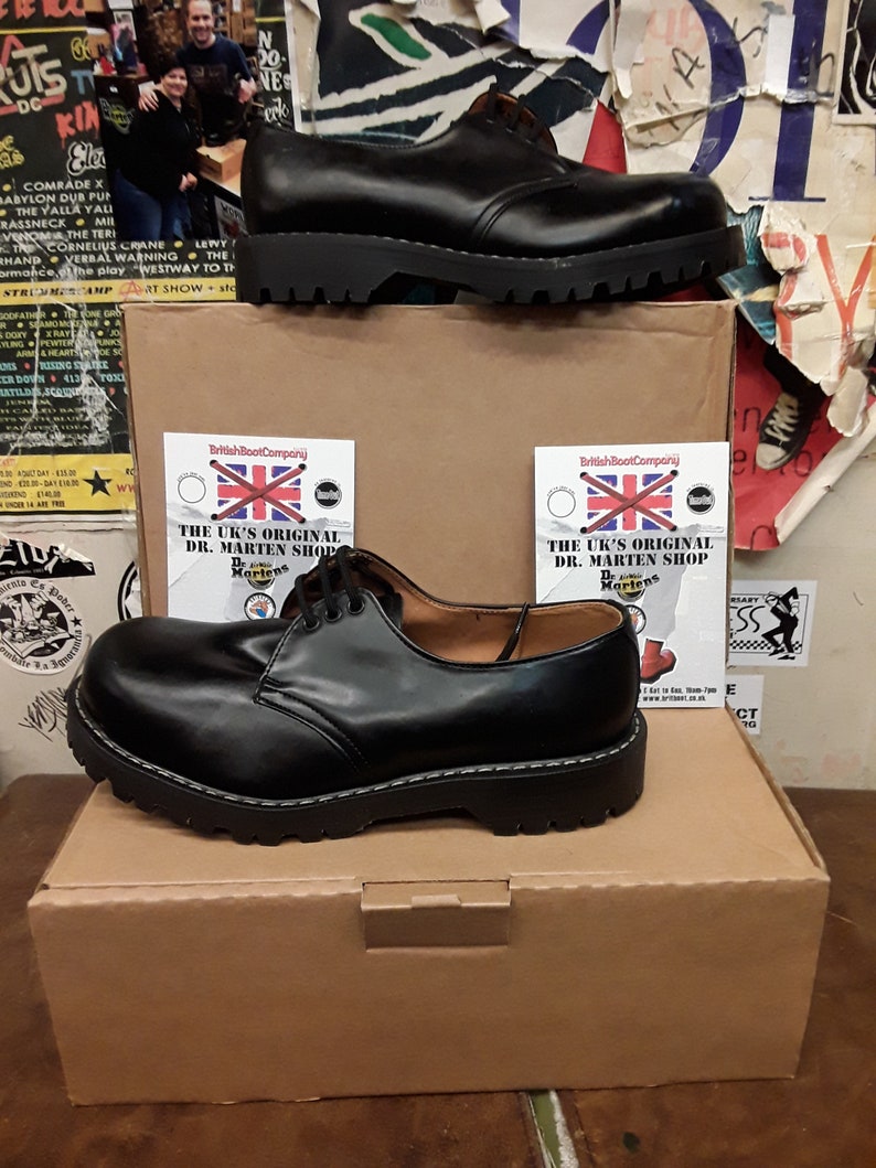 May include: A pair of black leather Dr. Martens shoes with a white stitching detail on the sole. The shoes are sitting on a brown cardboard box with the text "The UK's Original Dr. Martens Shop" printed on a white label.