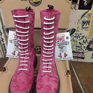 Puede incluir: Un par de botas Dr. Martens rosas con cordones blancos. Las botas están hechas de un material texturizado y tienen una suela negra.