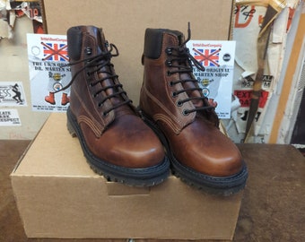 May include: A pair of brown leather boots with black laces and a thick black sole. The boots are sitting on a brown cardboard box. The box has a sticker on it that says "The UK's Original Dr. Martens Shop".