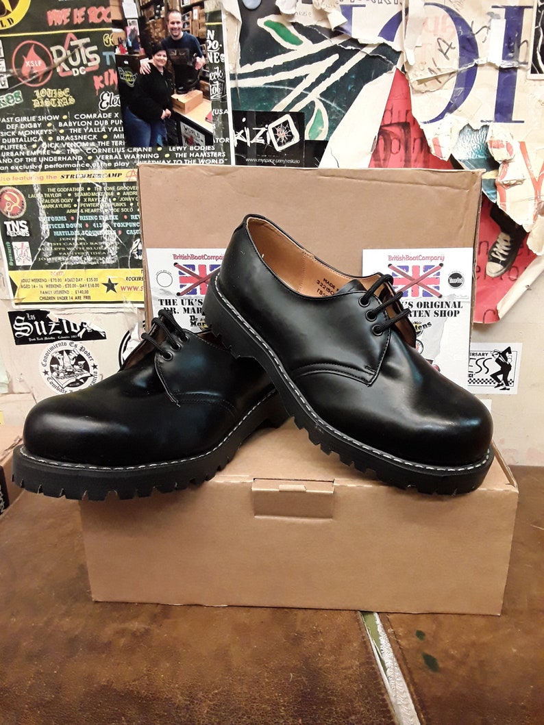 May include: A pair of black leather lace-up boots with a thick, black rubber sole. The boots are sitting on a brown cardboard box with the text "British Boot Company" and "The UK's Original Boot Shop" printed on it.