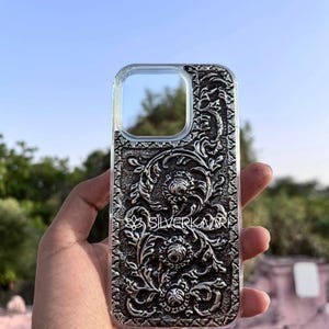 Puede incluir: Funda transparente para teléfono con un diseño floral plateado y ornamentado. La funda tiene forma rectangular con esquinas redondeadas y un recorte para la cámara. El texto "SILVERKARI" es visible.
