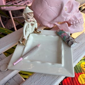 May include: A white ceramic square ashtray with a scalloped edge, holding a pre-rolled joint with a pink filter. A decorative elf figurine and a floral-patterned lighter are also present. A pink skull is in the background.