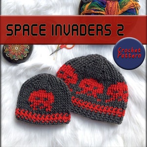 May include: Two crocheted hats in grey and red with a space invader design. The hats are on a white fluffy surface. The text "Space Invaders 2 Crochet Pattern" is on a red banner above the hats.