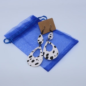 May include: A pair of dangle earrings with a black and white dalmatian print design. The earrings are made of wood and have a teardrop shape. The earrings are packaged in a blue organza bag.