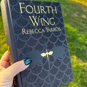 May include: A hardcover book titled "Fourth Wing" by Rebecca Yarros. The book has a black cover with a glittery, scale-like pattern and gold lettering. A gold dragon graphic is featured on the cover.
