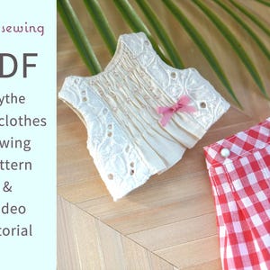 May include: A doll-sized cream-colored top with eyelet lace detailing and a pink bow, paired with red and white checkered shorts. The image also includes text that reads "Rosysewing PDF Blythe doll clothes sewing pattern & Video tutorial."