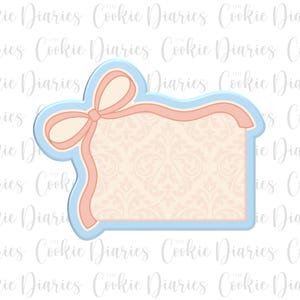 May include: A cookie cutter in the shape of a rectangular tag with a bow. The tag has a light blue border, a peach-colored bow and trim, and a cream-colored interior with a floral pattern. The text "Cookie Diaries" is in the background.