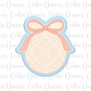 May include: A cookie cutter in the shape of a bow with a ribbon, a light blue outline, and a cream-colored interior with a floral pattern. The text "Cookie Diaries" is visible in the background.