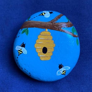 May include: A hand-painted, oval-shaped rock with a bright blue background. The rock features a yellow beehive, a brown branch, green leaves, and three bees. The bees are black and yellow with white wings. The rock is a decorative item.