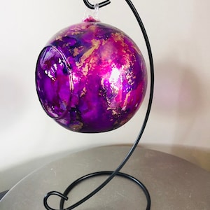 May include: A purple and gold glass bauble with a swirling pattern hangs from a black metal stand. The bauble is round and has a shiny finish.