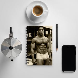May include: A sepia-toned spiral notebook featuring a muscular man in white briefs. A cup of coffee on a saucer, a black pencil, a silver coffee maker, and a black smartphone are arranged on a white surface.
