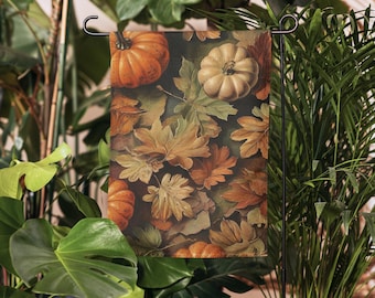 May include: A garden flag featuring a fall-themed design. The flag displays pumpkins in shades of orange and cream, scattered among fallen leaves in hues of brown, gold, and green. The flag is suspended from a black metal garden stake, surrounded by lush green foliage.