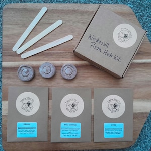 May include: A Windowsill Pizza Herb Kit by Pippin Seed Co. includes a cardboard box, three wooden plant markers, three compressed soil discs, and three brown seed packets labelled Oregano, Basil Genovese, and Chives with blue stickers.