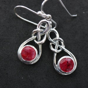 Può includere: Orecchini d'argento con un designo a nodo celtico e accenti di pietre preziose rosse.