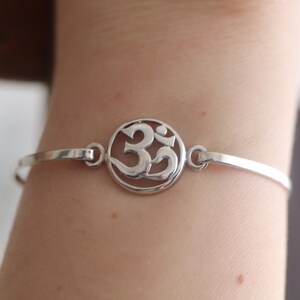 Puede incluir: Un brazalete de plata con un colgante circular que presenta un símbolo Om.