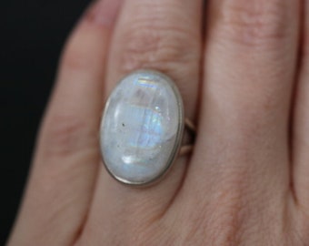 Puede incluir: Un anillo de plata con una gran piedra de luna ovalada. La piedra de luna es iridiscente y tiene un color blanco lechoso.