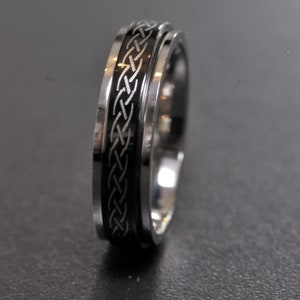 Op de afbeelding: Een zilverkleurige ring met een zwarte band met een Keltisch knoopontwerp. De ring heeft een gepolijste afwerking en een gedetailleerd, verweven patroon. De ring is een sieraad.