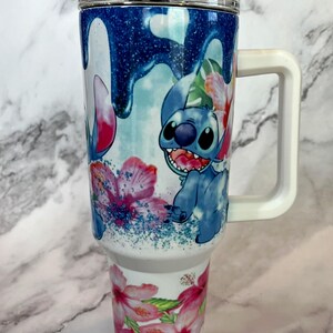 May include: White travel mug with a blue and white cartoon character design. The character is Stitch from Lilo & Stitch. The mug has a white handle and a clear lid. The design features pink flowers and blue glitter.